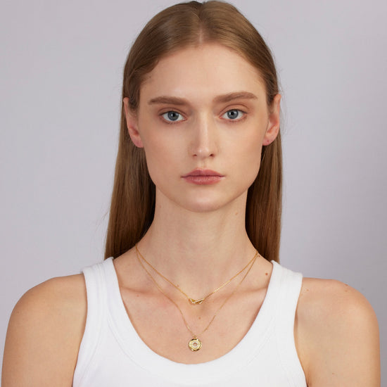 Woman wearing a white tank top and gold necklace against a plain background