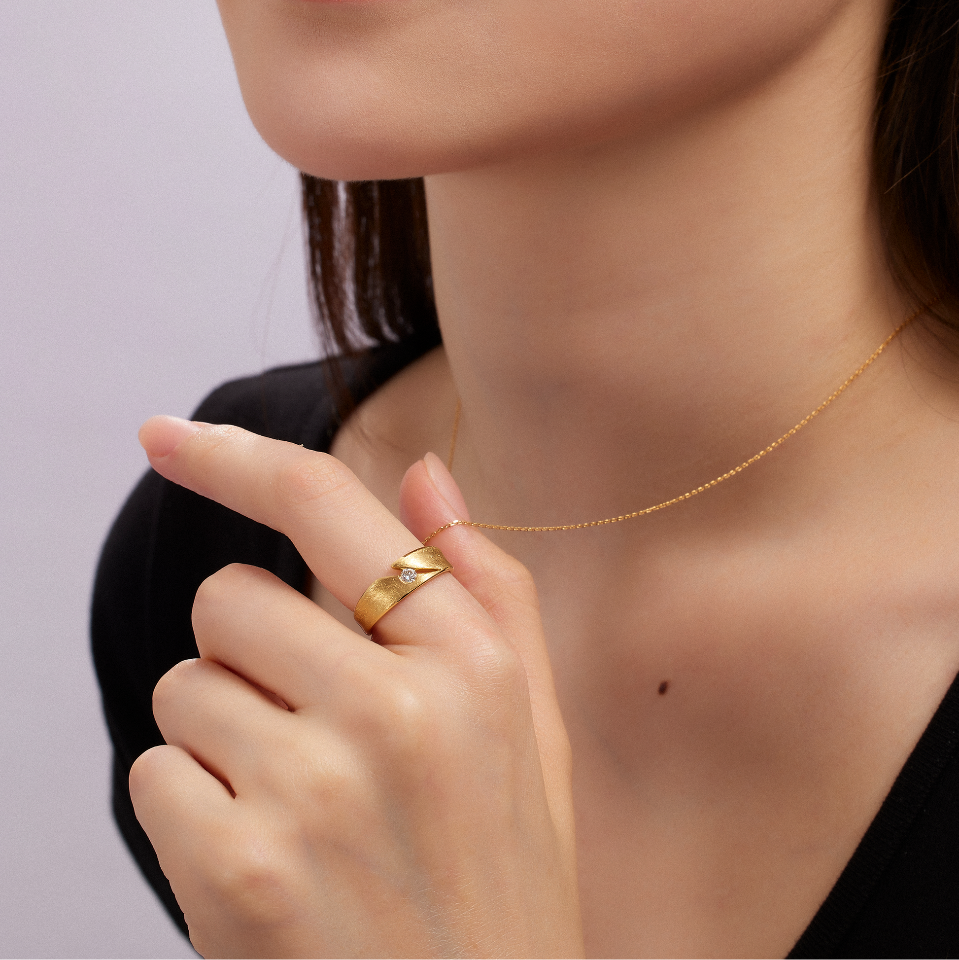 Close-up of a person wearing a gold necklace and ring against a light purple background