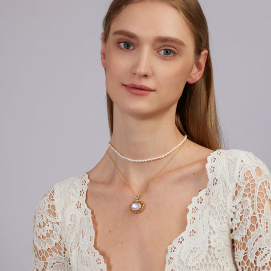 Woman wearing a white lace top and multiple necklaces on a plain background