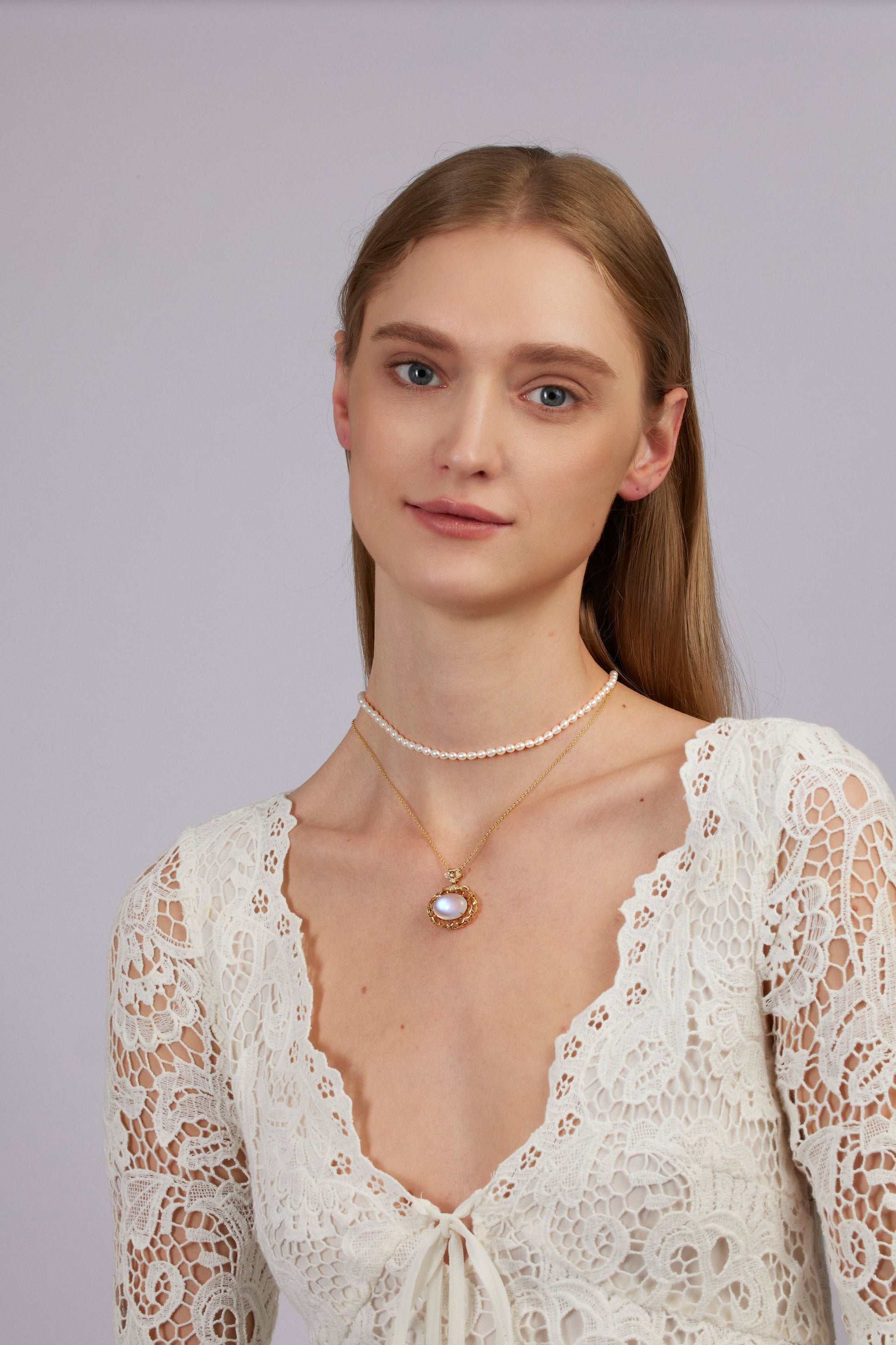 Woman wearing a white lace top and multiple necklaces on a plain background