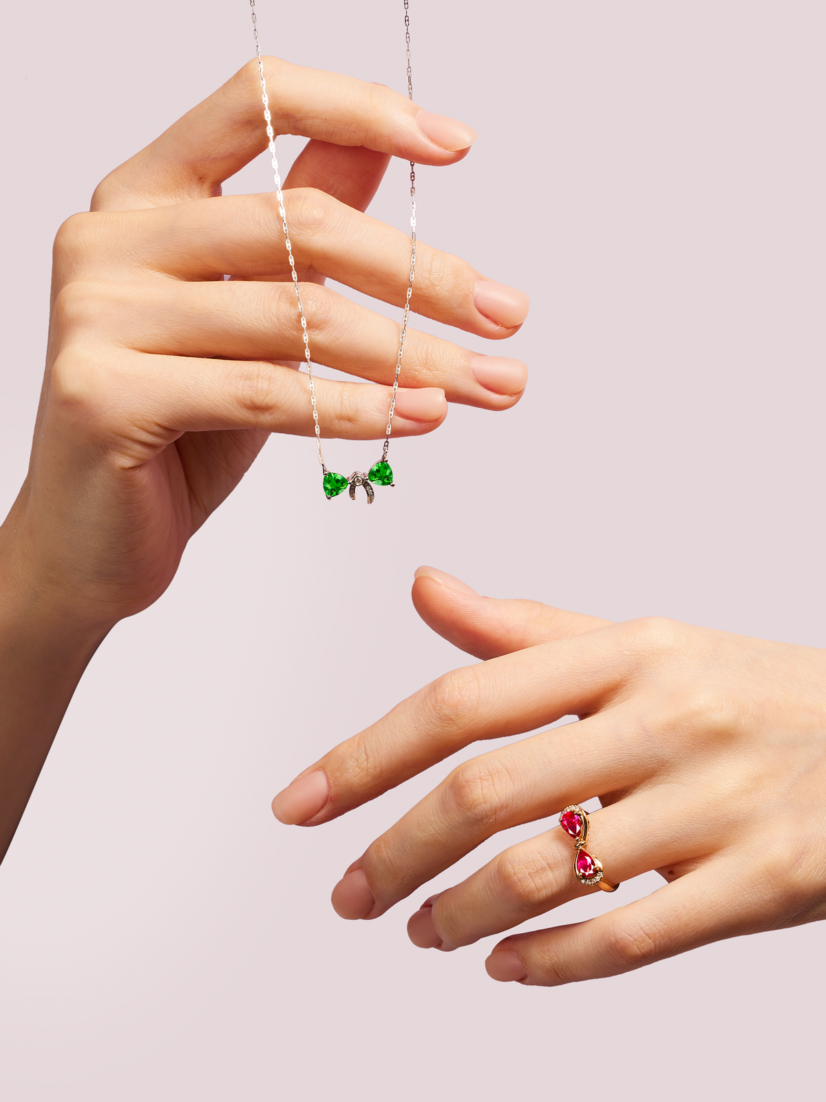 Hand holding a necklace with green gemstones and another hand wearing a ring with red gemstones on a light pink background.